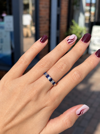A close-up of a hand wearing a slim white gold band ring with alternating dark blue and clear stones, set against a blurred outdoor storefront background.