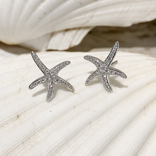 A pair of 18ct white gold starfish stud earrings set with small diamonds, displayed on a white seashell.
