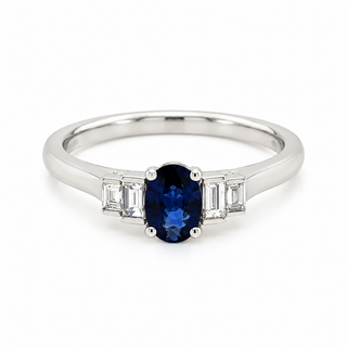 A silver band ring featuring an oval-cut deep blue sapphire center stone, flanked by two baguette-cut clear stones on each side, set against a plain white background.