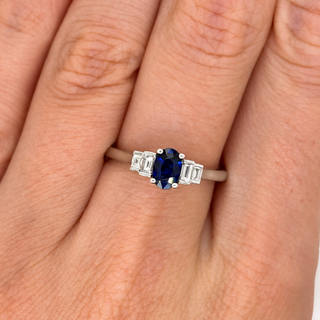 A close-up of a hand wearing a silver ring featuring an oval-cut deep blue sapphire center stone and baguette-cut side stones.
