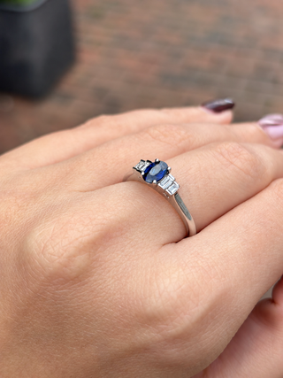 A silver ring with a blue oval center stone and two baguette-cut side stones, shown worn on a person's hand.