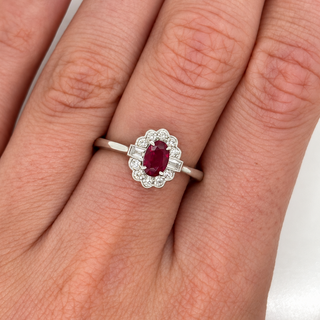 An 18ct white gold ring with an oval-cut red ruby center stone and a halo of small clear stones, shown worn on a person's finger.