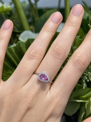 Hand wearing a ring with a pink gemstone against a green leafy background