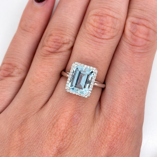A silver ring featuring an emerald-cut light blue aquamrine gemstone surrounded by a halo of small clear stones, shown worn on a person's finger.