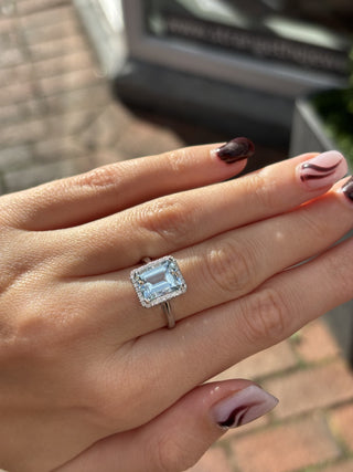 A close-up of a hand wearing an 18ct white gold ring with a rectangular emerald-cut light blue aquamarine center stone and a halo of small clear stones, photographed outdoors with a blurred brick-paved background.