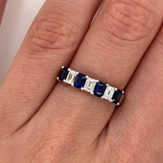 A close-up of a hand wearing a silver eternity ring set with baguette-cut deep blue sapphires and round-cut clear stones.