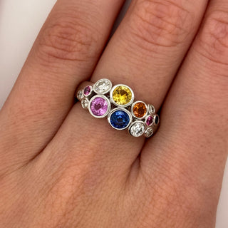 A close-up of a hand wearing a silver band ring set with multiple round, colorful gemstones in pink, blue, yellow, orange, and clear stones.
