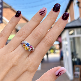 A close-up of a hand wearing a silver band ring adorned with multiple round-cut gemstones in yellow, blue, pink, and clear settings, photographed outdoors against a blurred street background.
