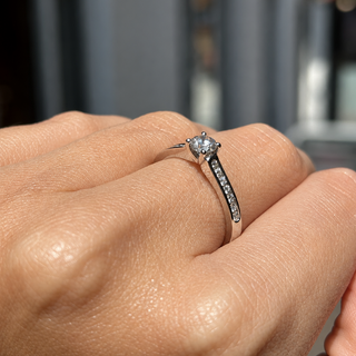A silver-toned diamond engagement ring with a round-cut center stone and a pavé-set diamond band, shown worn on a person's finger against a blurred outdoor background.