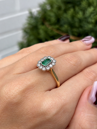 A close-up of a hand wearing a vintage-style ring with a green rectangular emerald-cut center stone surrounded by a halo of small clear stones, set on a yellow gold band.