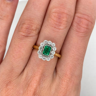 A close-up of a hand wearing a vintage-style emerald-cut green gemstone ring with a diamond halo and a yellow gold band.