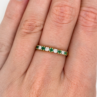 A close-up of a yellow gold eternity ring with alternating round-cut green emeralds and clear diamonds, worn on a person's ring finger.