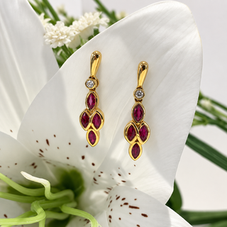 A pair of gold dangle earrings with marquise-cut ruby red gemstones and a small clear stone, displayed on the white petals of a lily of the valley.