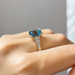 A close-up of a hand wearing a yellow gold ring featuring a rectangular emerald-cut blue gemstone and a pavé-set diamond band, set against a neutral gray background.