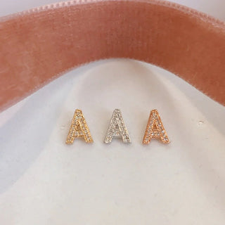 Three letter 'A' earrings in gold, silver, and rose gold on a white and pink fabric background.