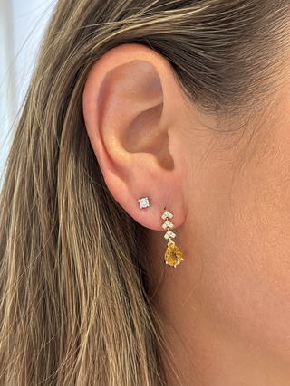 A close-up of a woman's ear wearing a small square stud earring and a gold drop earring with clear stones and a yellow pear-cut gemstone.