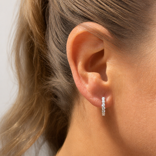 A close-up of a model's ear wearing a small silver huggie hoop earring with clear stones, with her hair tied back and a delicate gold necklace visible.