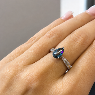 A close-up of a hand wearing a silver ring with a teardrop-shaped iridescent blue gemstone and small pavé-set stones on the band.