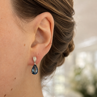A close-up side profile of a woman wearing a single teardrop-shaped blue gemstone dangle earring with a silver-toned setting and small clear accent stones.