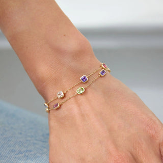 Hand wearing a gold bracelet with gemstones and a ring with a purple gemstone on a light background.