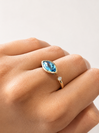 A close-up of a hand wearing a gold ring with a marquise-cut light blue gemstone, with a smaller diamond accent on the band, set against a clean off-white background.