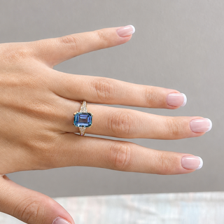 A close-up of a hand wearing a ring with a rectangular blue gemstone and a silver-toned band, set against a neutral gray background.