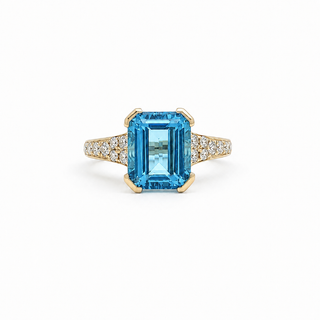 An emerald-cut blue topaz ring in a yellow gold setting, accented with pavé diamonds along the shoulders, shown on a clean white background.