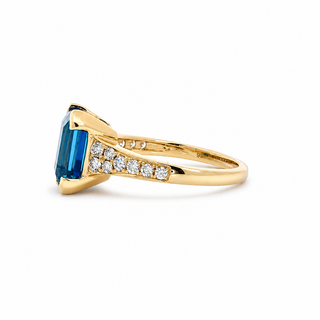 A yellow gold ring featuring a large emerald-cut blue gemstone and a row of small clear stones along the band, shown on a clean white background.