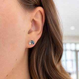 A close-up of a woman's ear wearing a small stud earring with a blue-green marquise-cut gemstone in a silver setting, against a softly blurred indoor background.