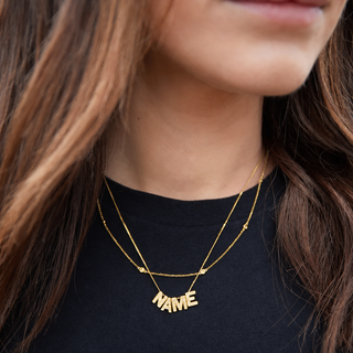 Gold 'NAME' necklace on a person wearing a black top