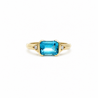 A yellow gold ring featuring a rectangular emerald-cut blue topaz center stone and small round diamonds on either side of the band, set against a plain white background.