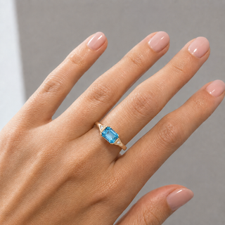 A close-up of a hand wearing a gold ring with an emerald-cut light blue gemstone, set against a neutral gray background.
