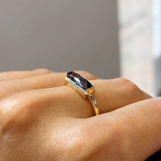 A close-up of a hand wearing a gold ring with a marquise-cut dark gemstone and small clear accent stones.