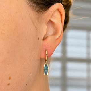 A close-up side view of a person's ear wearing a small gold hoop earring with a rectangular blue-green stone and a border of tiny clear stones, against a softly blurred window background.