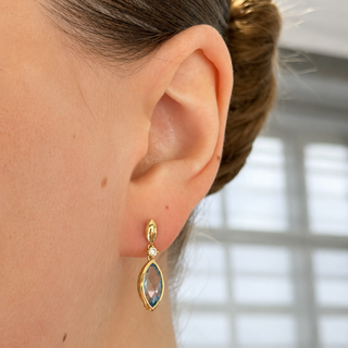 A close-up side view of a woman's ear wearing a gold drop earring with a marquise-shaped iridescent stone and a small clear accent stone.