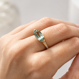 A close-up of a hand wearing a gold ring with a marquise-cut light green gemstone and small side stones, set against a soft, blurred light gray background.