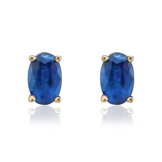 A pair of oval-cut blue gemstone stud earrings in a gold four-prong setting, displayed on a clean white background with soft reflections below.
