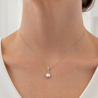 A close-up of a model's neck and upper chest wearing a delicate gold chain necklace with a white pearl pendant and small clear accent stones.