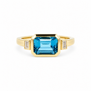 A yellow gold ring featuring a large rectangular emerald-cut blue topaz center stone, accented by two small baguette-cut diamonds on either side of the band, set against a plain white background.