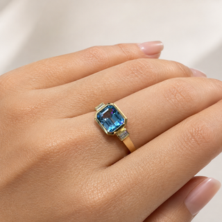 A gold ring with a rectangular blue gemstone, worn on a person's finger against a soft, light background.