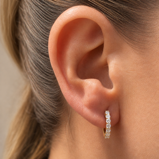 A close-up of a model's ear wearing a small gold huggie hoop earring set with clear pavé gemstones.
