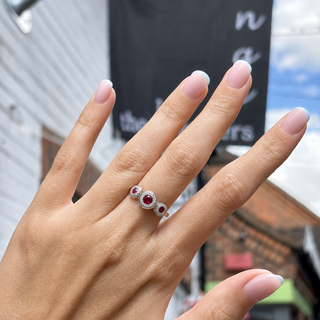 A close-up of a hand wearing a silver ring with three oval-cut red gemstones, set against a blurred outdoor background of a building and sky.