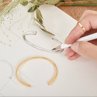 Hand drawing a gold bracelet on paper with a pencil, surrounded by greenery.