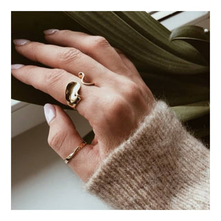 Hand wearing a gold ring with a leaf design, holding a phone, against a blurred indoor background.