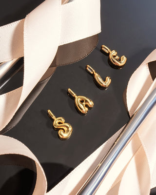 Four gold initial pendant charms spelling 'SCSC' arranged on a black surface with beige ribbon accents.
