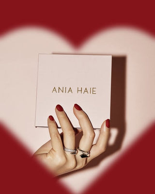 Hand holding a pink 'Ania Haie' box with red heart-shaped cutout in the background