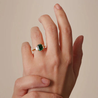 Hand wearing a gold ring with a green gemstone on a beige background