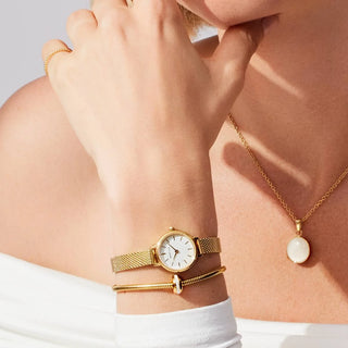 Gold watch and bracelets on a wrist with a gold necklace on a neutral background
