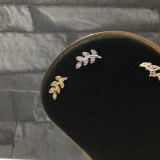 A pair of leaf-shaped stud earrings displayed on a black jewelry stand. One earring is gold-toned with clear stones, and the other is silver-toned with clear stones.
