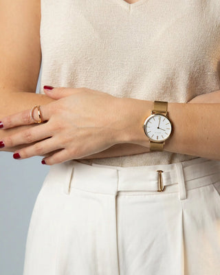 A close-up of a person wearing a slim gold-tone wristwatch with a white dial and matching gold-tone mesh strap, paired with a beige knit top and white trousers.